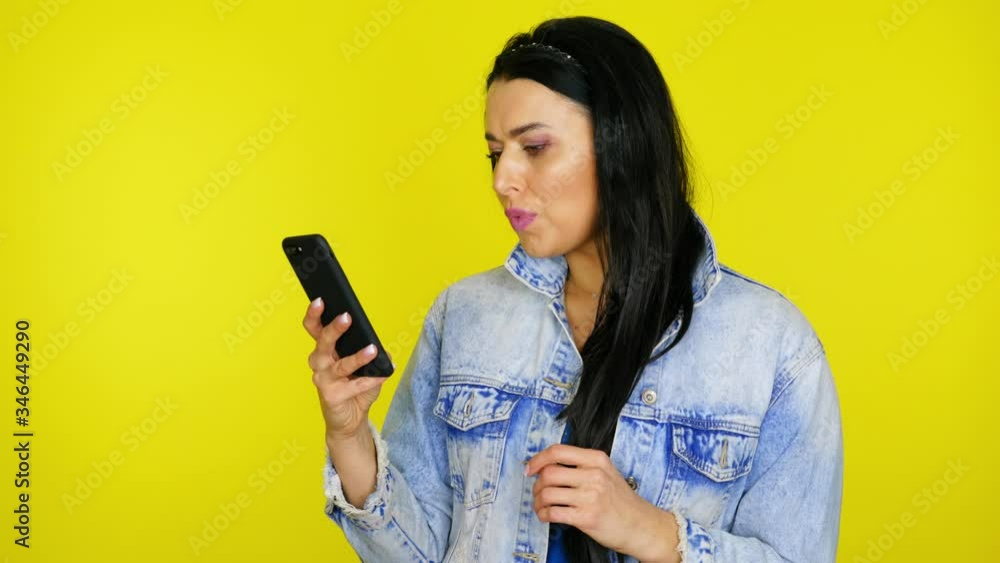 Woman uses a smartphone then is surprised and says wow on yellow background with copy space. Girl with black hair in a denim jacket is looking for discounts in online stores. Place for text or product Woman uses a smartphone then is surprised and says wow on yellow background with copy space. Girl with black hair in a denim jacket is looking for discounts in online stores. Place for text or product