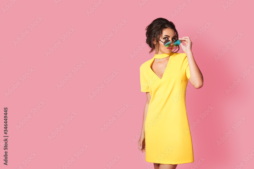 Fototapeta premium Beautiful girl wearing yellow dress and sunglasses posing on pink background in studio. Looking at camera.