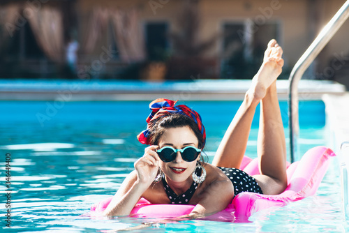  Beautiful girl by the pool with long dark hair and make-up resting at a spa resort. In a black retro bathing suit, scarf, hat and glasses, smiles sunbathes on a pink inflatable mattress in the motel