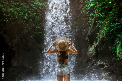 Beautiful young sexy tourist girl with straw hat from back in the tropical jungle looks at a strong and rapid waterfall. Adventure, tourism, exploration and travel concept.