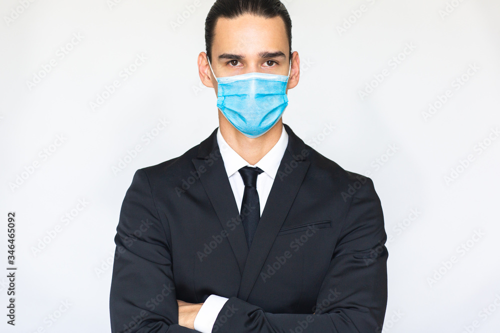 Confident young businessman in suit with arms crossed wearing a blue anti virus medical mask and looking at camera. Isolated portrait concept on white background. Confident young businessman in suit with arms crossed wearing a blue anti virus medical mask and looking at camera. Isolated portrait concept on white background.