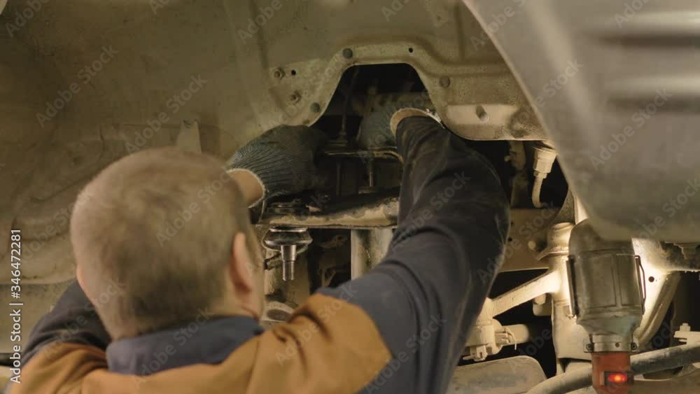 An Asian man is repairing the chassis of an old car lifted on a lift in a workshop, close-up. An Asian man is repairing the chassis of an old car lifted on a lift in a workshop, close-up.