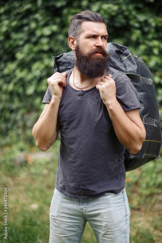 Brutal bearded male courier in shorts and a T-shirt with a big gray bag on his shoulders in a green park. homeless not shaved man with a big gray bag.
