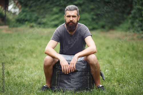 Brutal bearded male courier in shorts and a T-shirt with a big gray bag is resting in a green park. homeless man not shaved with things in nature.