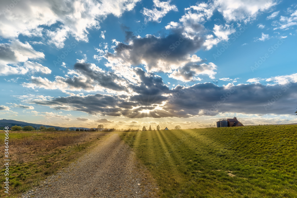 An imposing cloud sky in a landscape of fields and a path where sun rays come out of the holes