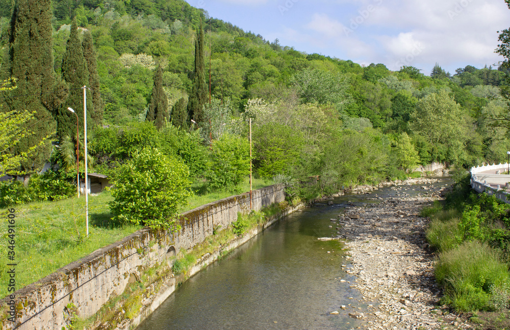 Spring landscape. A mountain meandering river enclosed in stone fences. Shallow, with a bare stone bottom on the side. Green plants, shrubs, and trees. Sunny weather. Spring landscape. A mountain meandering river enclosed in stone fences. Shallow, with a bare stone bottom on the side. Green plants, shrubs, and trees. Sunny weather.