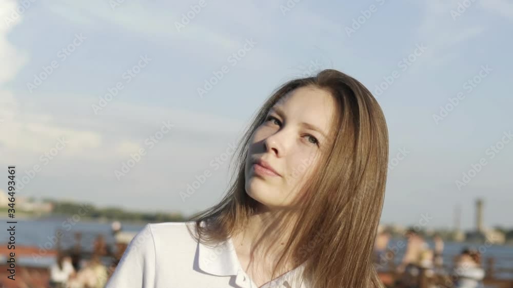 Slow motion of portrait of young blond woman looking into camera, cute smile, sun shining brightly, river background. Cute face model smiling. Girl looks joy at camera. Blond hair fly fluttering wind Slow motion of portrait of young blond woman looking into camera, cute smile, sun shining brightly, river background. Cute face model smiling. Girl looks joy at camera. Blond hair fly fluttering wind