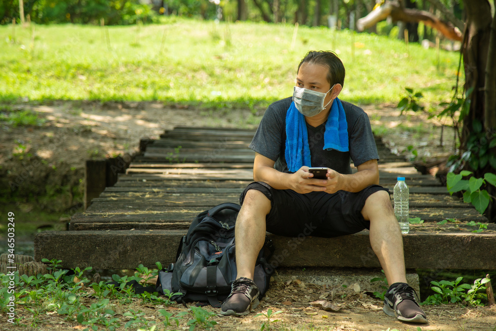 Asian man in black casual wear wearing a flu mask sitting on old boards of a wooden bridge in public park with grreen lawn nature background Asian man in black casual wear wearing a flu mask sitting on old boards of a wooden bridge in public park with grreen lawn nature background