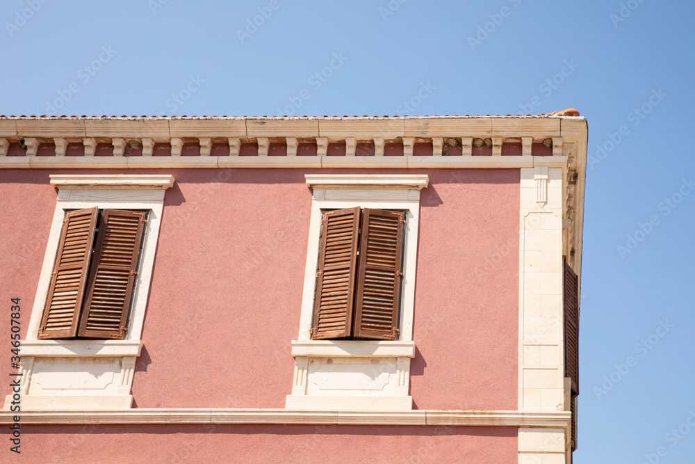 Close up of the exterior of a old pink colored house with white details and wooden louvre windows half closed. Typical mediterranean style. On a sunny day with a blue sky.