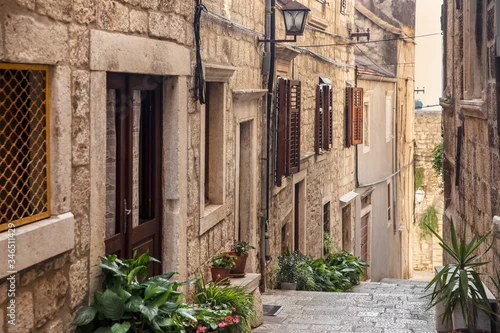 Obraz Korcula old narrow Mediterranean street with stairs. Rough stone houses, facades with windows, green plants, flowers in Dalmatia, Croatia. Historical place creating a picturesque and idyllic scenery