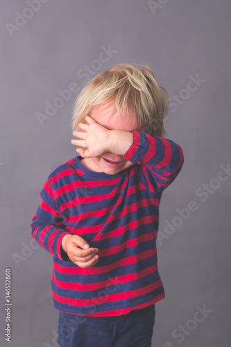 Little toddler child, boy, crying, hiding his face with hand, isolated image on gray