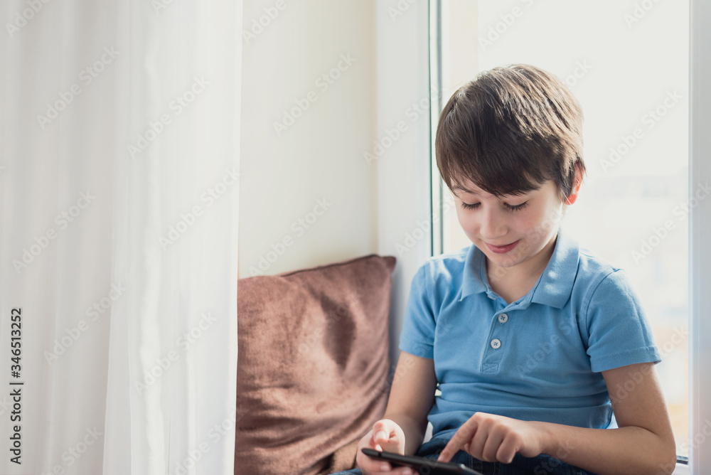 Boy playing with tablet sitting on pillow and windowsill. Soft focus. Quarantine and online school concept