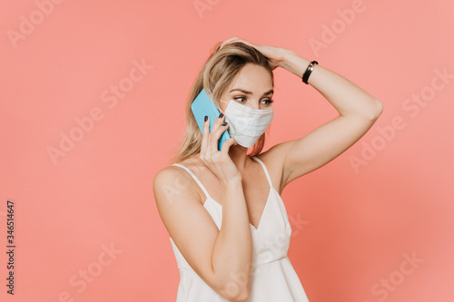 Beautiful blonde in protective mask speaking by cellphone with her aged parents  she is worried about their state because of raising pandemic of coronavirus. Over pink backdrop. Healthcare concept.
