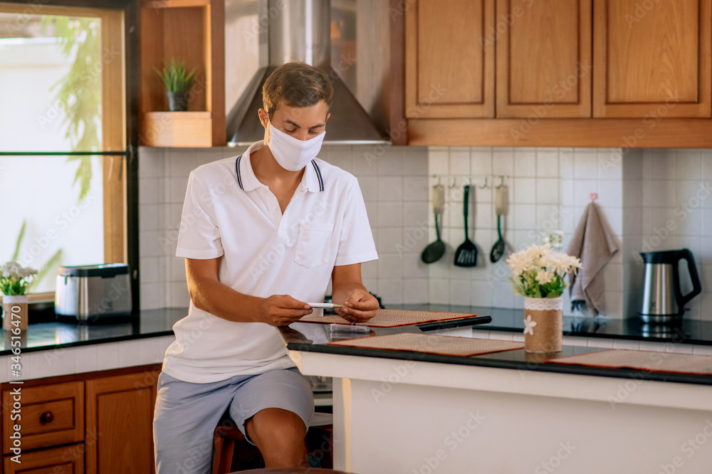 A young man in a white protective medical mask takes a thermometer from its case to measure the temperature of his body. A man is sitting in the kitchen of his house. Quarantine. A young man in a white protective medical mask takes a thermometer from its case to measure the temperature of his body. A man is sitting in the kitchen of his house. Quarantine.