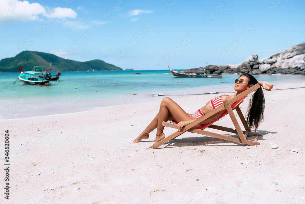 Beautiful girl on a lounger in a striped red and white bikini. Asian brunette relaxing on the beach. Tropical vacation. Thai boats and blue sea on the background Beautiful girl on a lounger in a striped red and white bikini. Asian brunette relaxing on the beach. Tropical vacation. Thai boats and blue sea on the background