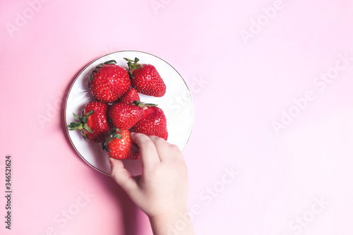 Top view Childs hand holding a strawberry on pink background, plate of strawberries. Summer healthy eating concept.
