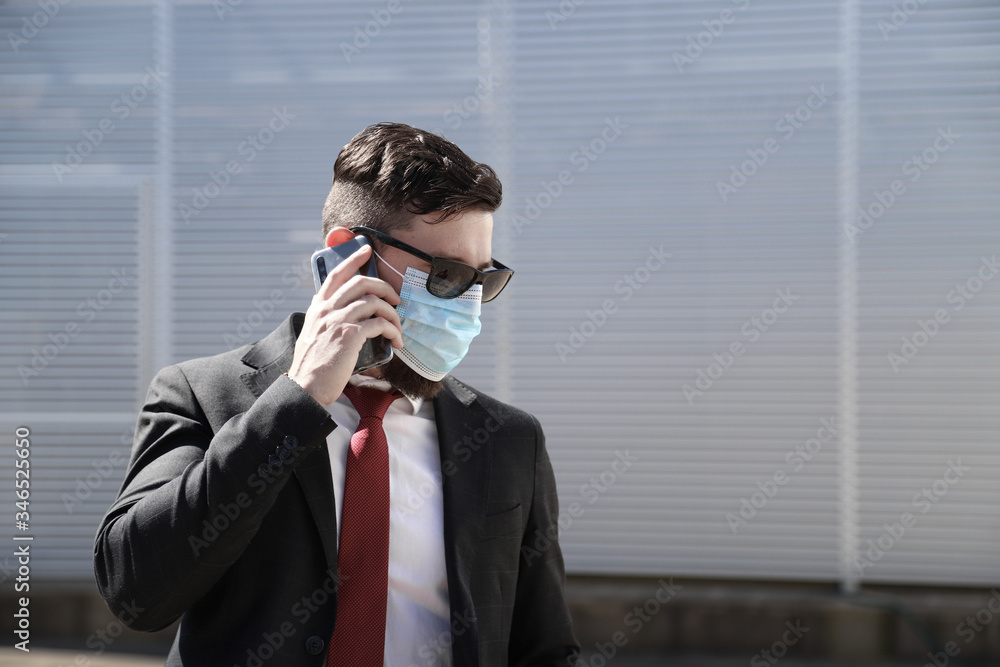 Businessman with hipster look wearing surgical face mask talking on his smartphone for news updates of coronavirus outdoors. Modern architecture on the background Businessman with hipster look wearing surgical face mask talking on his smartphone for news updates of coronavirus outdoors. Modern architecture on the background