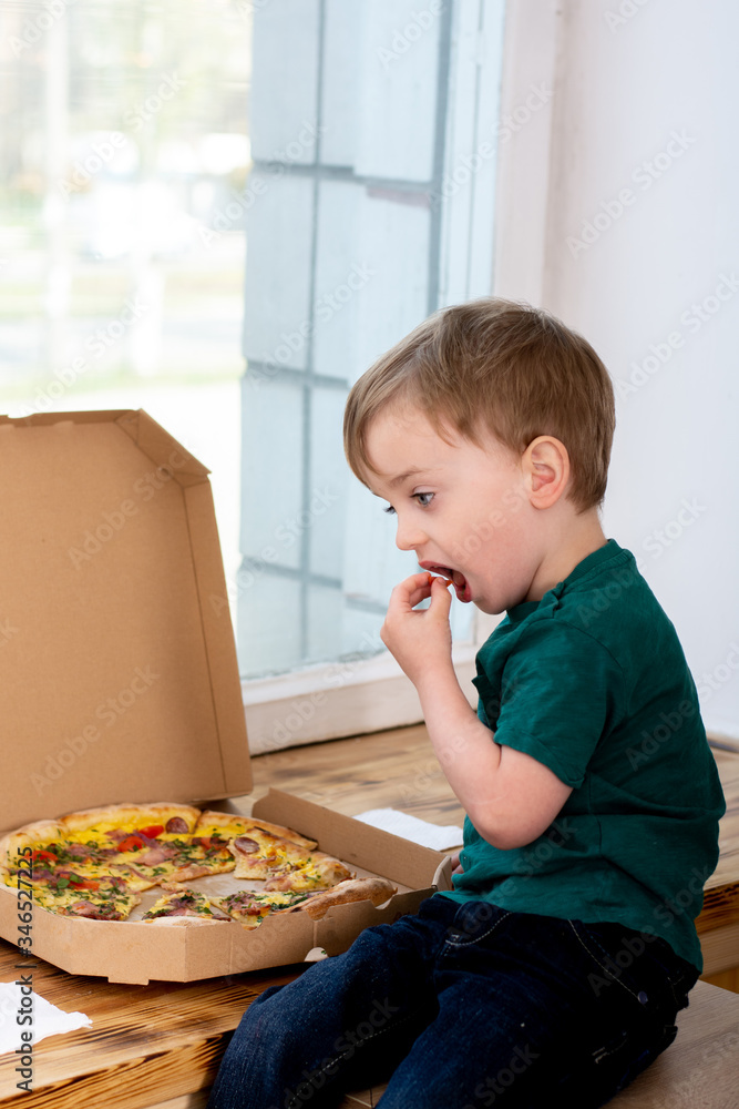 little hungry child eating a slice of pizza near the window in the apartment. the boy puts his hands a slice of tomato in his mouth. close-up. little hungry child eating a slice of pizza near the window in the apartment. the boy puts his hands a slice of tomato in his mouth. close-up.