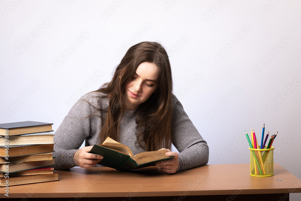 a young girl sits at a table and reads a book. student preparing for the exam. gray wall on the background. a jar with multi-colored pencils stands nearby. a young girl sits at a table and reads a book. student preparing for the exam. gray wall on the background. a jar with multi-colored pencils stands nearby.