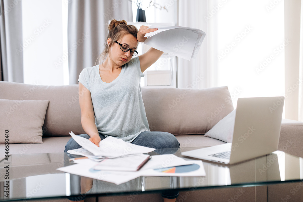 Tired young girl in home clothes works remotely at home. She works with paperwork and laptop while being at home. Distant work Tired young girl in home clothes works remotely at home. She works with paperwork and laptop while being at home. Distant work