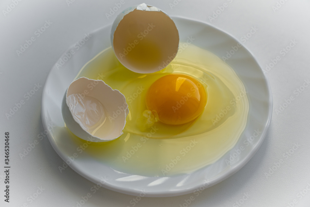 One cracked egg with yellow yolk surrounded by egg shells is laying on a white plate on white background One cracked egg with yellow yolk surrounded by egg shells is laying on a white plate on white background