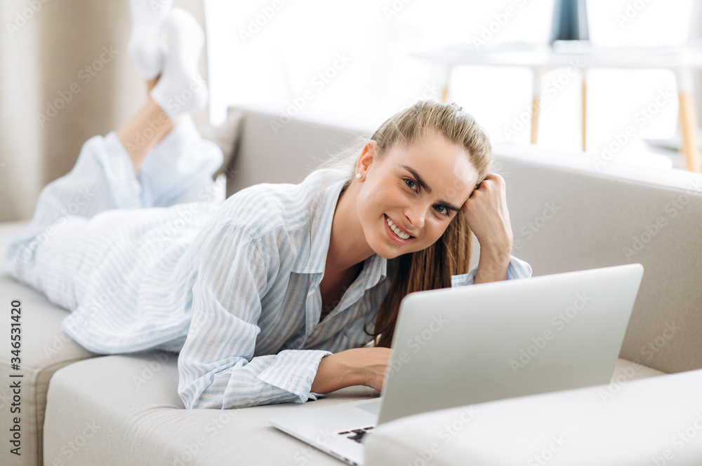 Weekend morning. A young amazing girl in pajamas lying on the couch and spends time in network chatting with friends and shopping online  using a laptop