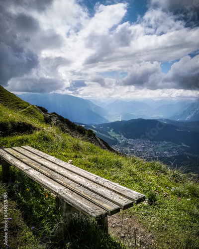 Dramatic alpine panoramic top view across valley with mountains and village in background, wooden bench close up