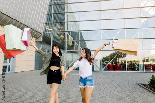 shopping and tourism concept - beautiful Asian girls with shopping bags  holding hands and walking together on the backgrund of modern urban shopping center outdoors