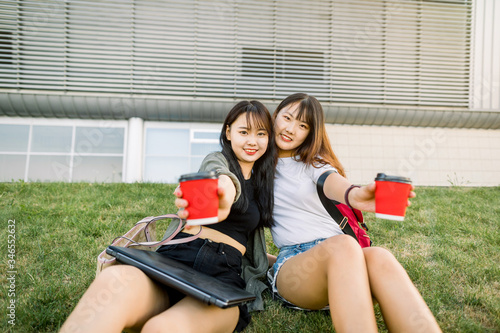 Beautiful Asian female college students holding cups of coffee and showing them to camera, sitting outdoor on green grass in front of modern urban building. People, friendship and leisure