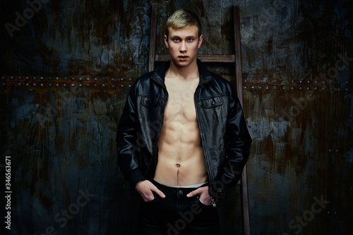 Handsome young shirtless man in leather jacket posing against dark studio background.
