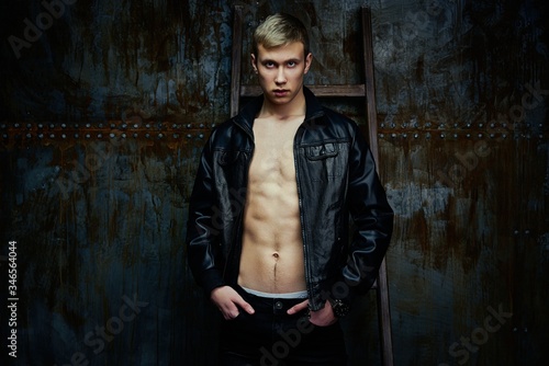 Handsome young shirtless man in leather jacket posing against dark studio background.