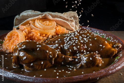 Exotic Mexican food on a mud or clay plate. Chicken Mole from Oaxaca with sesame seeds falling from above and a side of Mexican tomato rice and corn tortillas in a black background and a wooden table