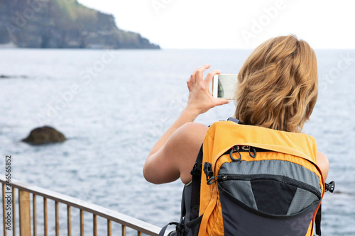 A red-haired girl with a backpack on her back takes photos of the ocean on her phone. Copy space.