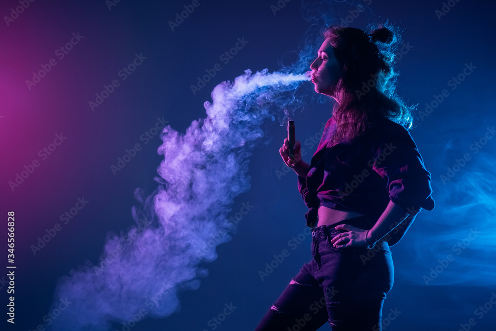 Smoking an electronic cigarette. The girl stands sideways and lets smoke out of her mouth. The concept of vaping. A woman and smoke from an e-cigarette on a dark background.