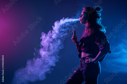 Smoking an electronic cigarette. The girl stands sideways and lets smoke out of her mouth. The concept of vaping. A woman and smoke from an e-cigarette on a dark background.