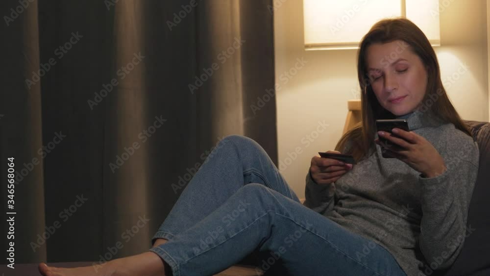 Woman lying on the couch in a cozy room and makes an online purchase using a credit card and her smartphone. Online shopping, lifestyle technology. Woman lying on the couch in a cozy room and makes an online purchase using a credit card and her smartphone. Online shopping, lifestyle technology.