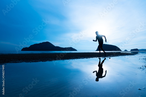 Silhouette Asian man running on the beach with shadow on water during sunrise toned in blue color
