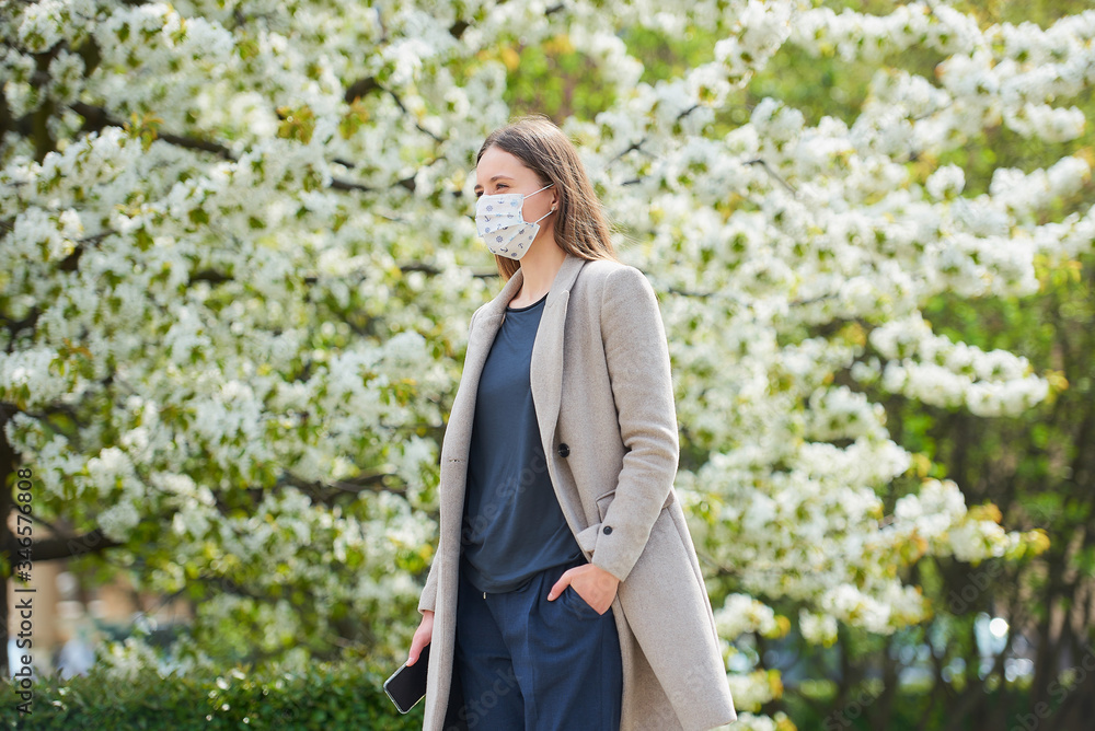 A girl in a medical face mask to avoid the spread coronavirus holds a smartphone in the park. A woman in a face mask against COVID-19 keeps a social distance in the garden between flowering trees.