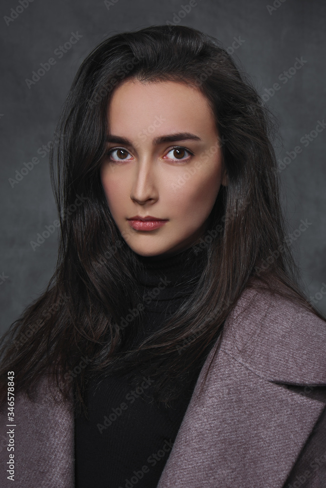 Beautiful young brunette girl in a black sweater and lilac coat on a gray background.