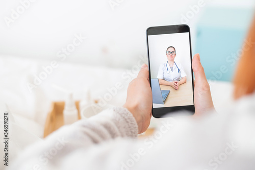 The concept of distance treatment. Rear view of a woman in bed in pajamas with a smartphone in her hands. A flu patient is watching a medical video blog.