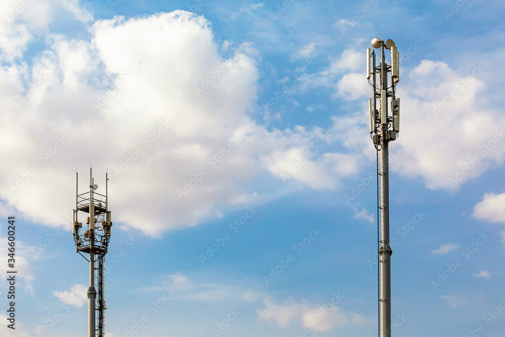 Two telecommunications towers with 4G, 5G transmitters. cellular base station with transmitting antennas on a telecommunications tower against a blue sky with clouds. Two telecommunications towers with 4G, 5G transmitters. cellular base station with transmitting antennas on a telecommunications tower against a blue sky with clouds.
