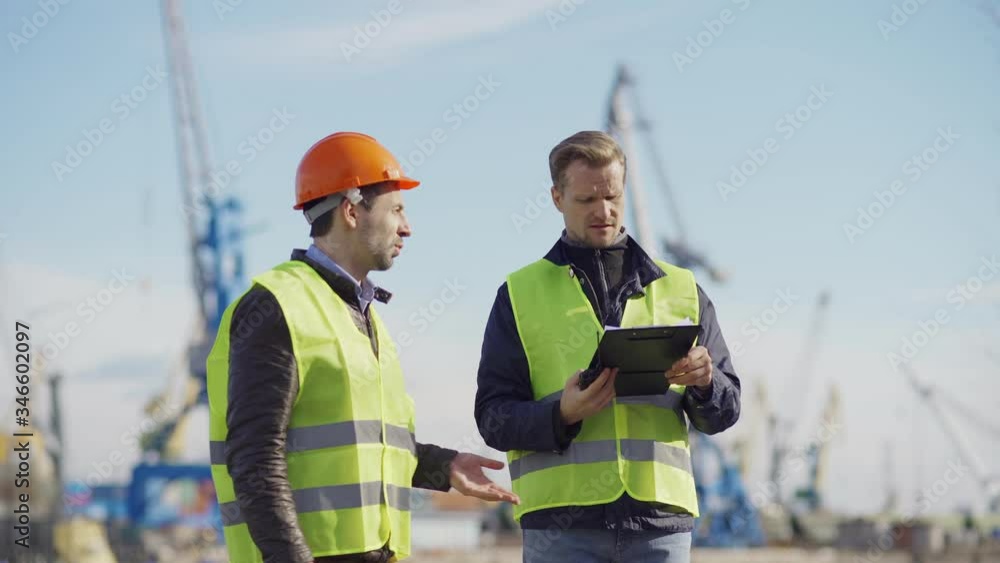 Medium shot of two engineers walking at shipbuilding yard, observing constructions of vessels and discussing plans. Middle aged specialist with clipboard speaking into walkie-talkie Medium shot of two engineers walking at shipbuilding yard, observing constructions of vessels and discussing plans. Middle aged specialist with clipboard speaking into walkie-talkie
