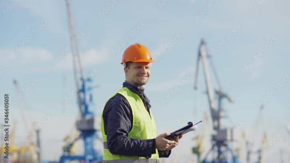 Side view medium shot of middle aged marine engineer in orange helmet and with clipboard speaking into walkie-talkie at dockside, then turning his head, looking at camera and smiling Side view medium shot of middle aged marine engineer in orange helmet and with clipboard speaking into walkie-talkie at dockside, then turning his head, looking at camera and smiling