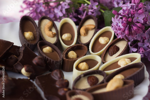 Bouquet of lilac flowers and chocolate candies with filling and nuts, chocolates with nuts on a plate, background. Sweets made of black, milk and white chocolate with hazelnuts, almonds and cashews