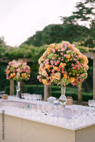 Wedding dinner table reception. Huge bouquets of yellow, pink, orange roses and green hydrangeas in a glass vase on a high leg. Large bouquets in the shape of a ball. A bar with lots of glasses.