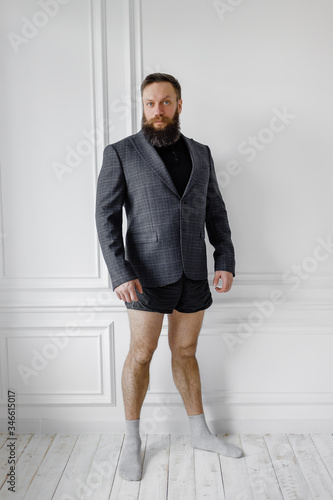 bearded man in a jacket and without pants stands against a light wall