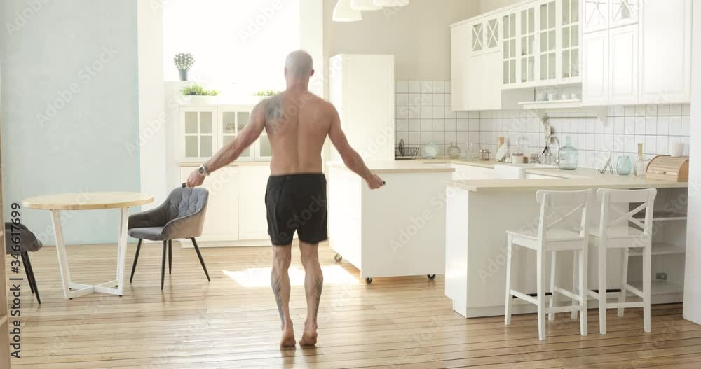 Muscular athletic bald man is jumping rope making cardio training at home in kitchen, back view. Skipping rope. Sport and fitness. Workout and wellness concept. High-intensity interval training. Muscular athletic bald man is jumping rope making cardio training at home in kitchen, back view. Skipping rope. Sport and fitness. Workout and wellness concept. High-intensity interval training.