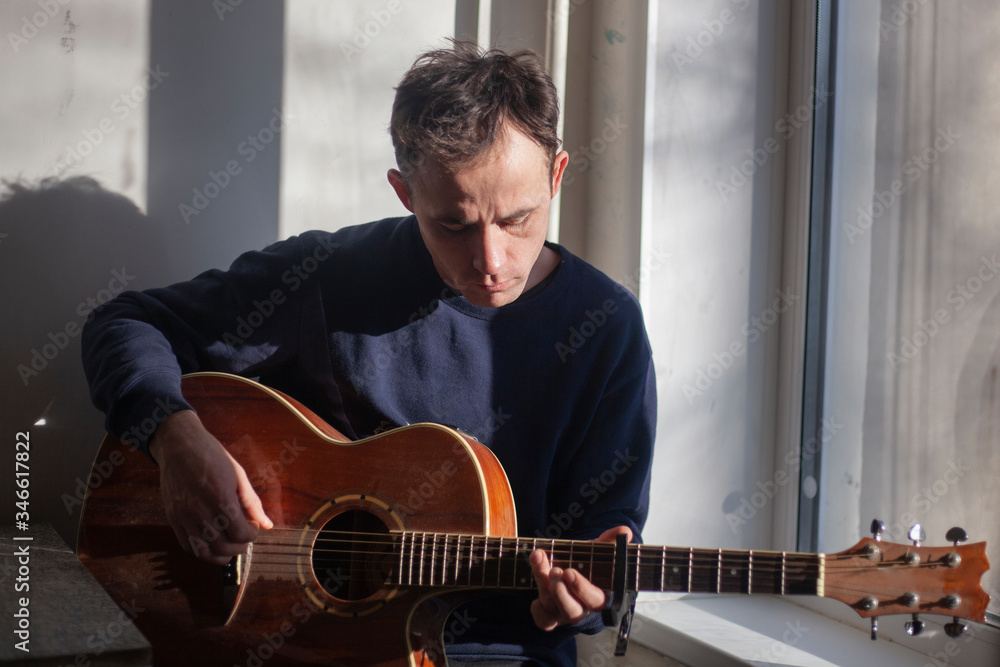 Obraz premium Guy with a guitar near the window. The guy in the morning light in the kitchen. The man is rehearsing music. Playing an acoustic instrument. Guitar lesson.