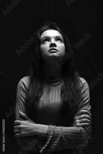 Black and white portrait of a young girl looking up girl with long hair.  Woman prying. Top light. Dramatic girl on a dark background. Selective lighting. Concept of religion, hope and prayers.