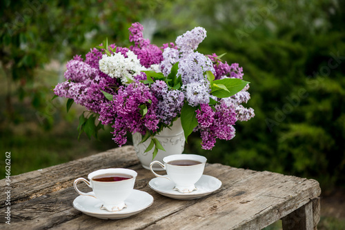 Lilac spring flowers in vase on wooden background. Tea coffee cup on table during breakfast time